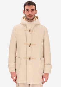 Uomo che indossa un cappotto toggole in corduroy beige con cappuccio, due grandi tasche frontali e cinturini alle maniche, in piedi contro uno sfondo semplice.
