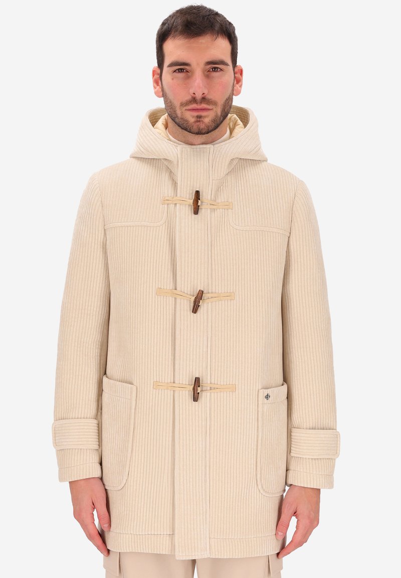 Uomo che indossa un cappotto toggole in corduroy beige con cappuccio, due grandi tasche frontali e cinturini alle maniche, in piedi contro uno sfondo semplice.