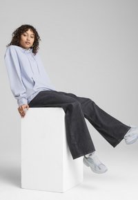Felpa azzurra chiara con tasca frontale, jeans neri a gamba larga e sneakers grigie con accentuazioni blu, seduto su un piedistallo bianco.