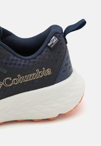 Pantofii sport impermeabili de culoare bleumarin au o parte superioară din plasă texturată, logo-ul auriu "Columbia" și o talpă albă cu perne de amortizare, dotată cu crampoane portocalii pentru aderență.