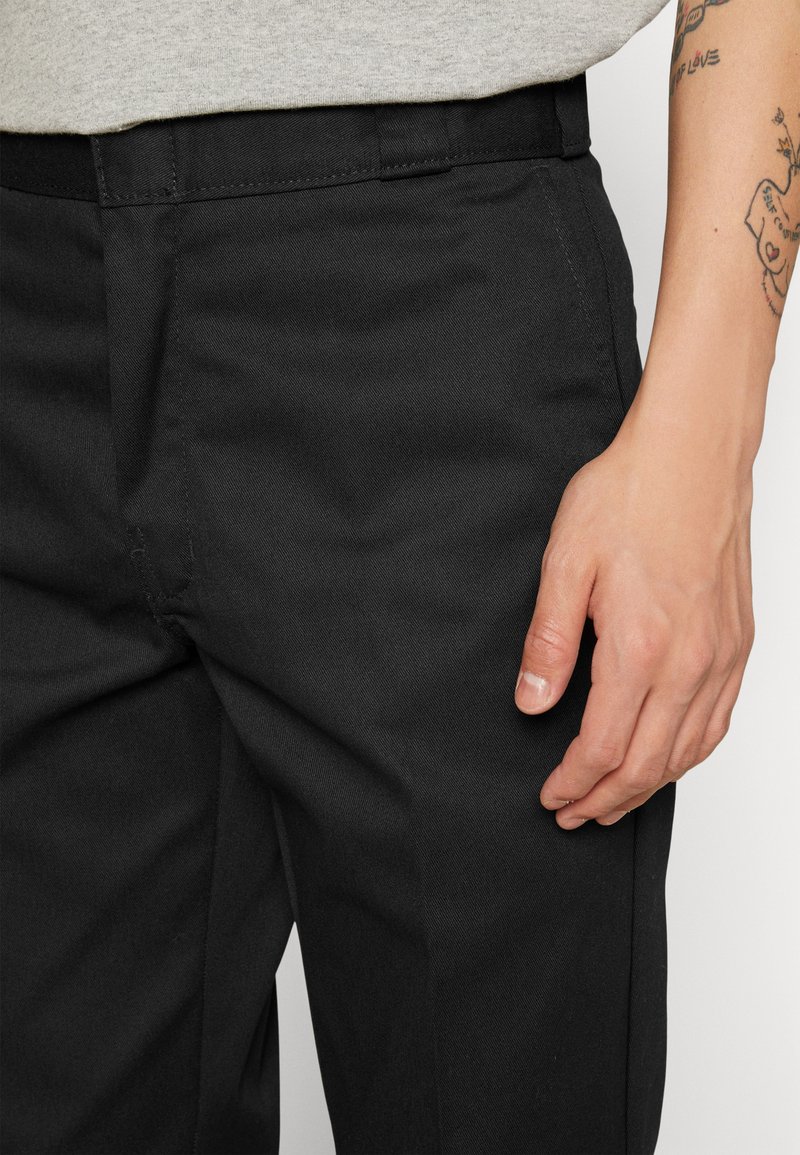 Black trousers made of a sturdy fabric, with a tailored fit, flat front, and side pockets. Visible hand resting on the fabric.