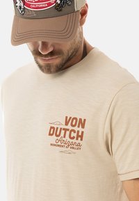 Camiseta de manga corta beige con un pequeño logo marrón que dice "VON DUTCH Arizona Monument Valley" y un gráfico de montañas en el frente.