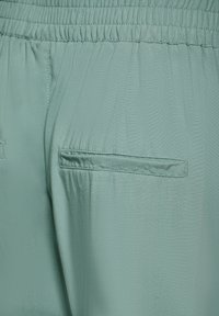 Světle zelené kalhoty s hladkým povrchem, vybavené elastickým pasem a dvěma zadními kapsami. Jednoduchý design bez viditelných vzorů.