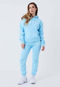 Felpa leggera blu e pantaloni jogger abbinati, realizzati in materiale morbido, con tasca frontale e ricamo del logo. Abbinati a sneakers bianche.