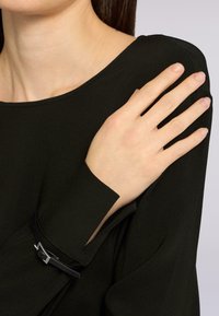 Persona con una maglietta nera a maniche lunghe caratterizzata da un piccolo dettaglio con cintura nera sul polsino, con la mano appoggiata sulla spalla opposta.