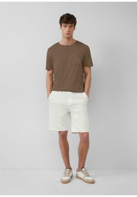 Braunes Kurzarm-T-Shirt mit Brusttasche, kombiniert mit weißen Streifen-Shorts und beigen Sneakern mit Gummisohlen.