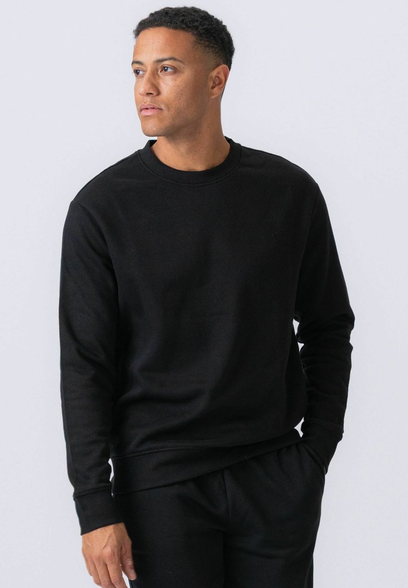 Mann mit kurzen lockigen Haaren, trägt schwarzen Pullover und Hose, steht mit einer Hand in der Tasche und blickt nach links auf einem schlichten hellen Hintergrund.