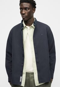 Νέος άντρας που φορά στρωμένα ρούχα με σκούρο μπλε πουκάμισο με κουμπιά, ανάγλυφο ανοιχτοπράσινο πουκάμισο, λευκό μπλουζάκι και πράσινο παντελόνι.