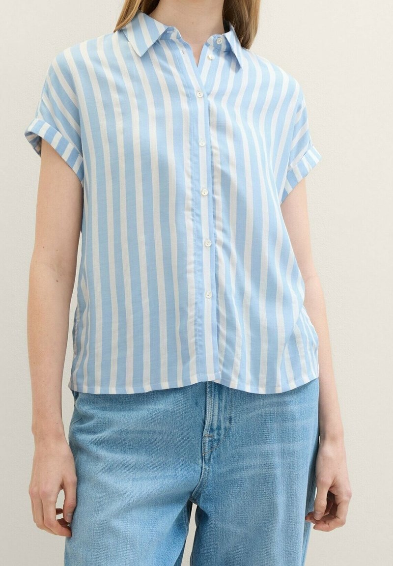 Personne portant une chemise boutonnée à manches courtes à rayures verticales bleu clair et blanc avec manches retroussées et un jean en denim bleu clair.