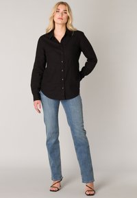 Zwarte button-up shirt met een zak, lange mouwen en een kraagontwerp, gecombineerd met lichtblauwe rechte jeans en zwart gekruiste sandalen.