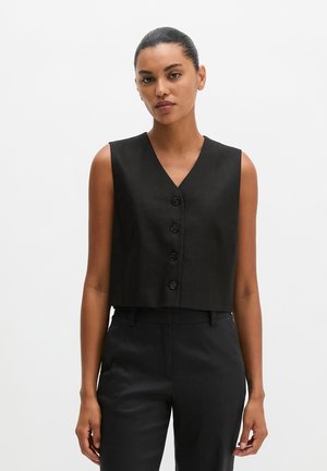 Femme aux cheveux foncés en chignon lisse portant un gilet noir sans manches boutonné et un pantalon noir taille haute assorti, debout devant un fond blanc.