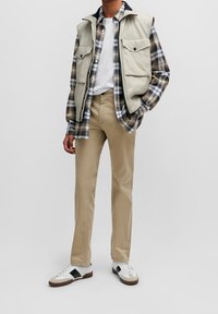 Veste cargo beige portée sur une chemise à carreaux bleu, marron et blanc ; t-shirt crème ; pantalon beige ; baskets blanches avec des accents noirs.