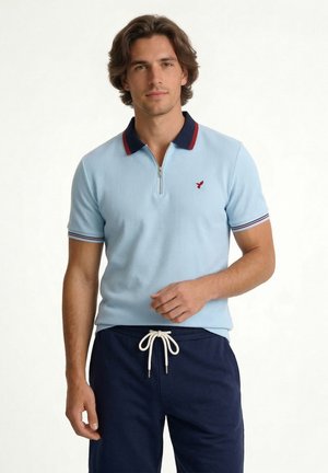 Mężczyzna ubrany w jasnoniebieskie polo z krótkim rękawem, granatowym i czerwonym kołnierzykiem, granatowe spodnie ze sznurkiem, stojący na jednolitym tle.