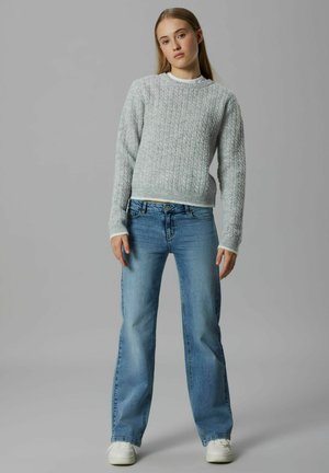 Šedý svetr z pleteniny s žebrovaným výstřihem, doplněný světle modrými džíny s volným střihem. Model stojí proti neutrálnímu pozadí.