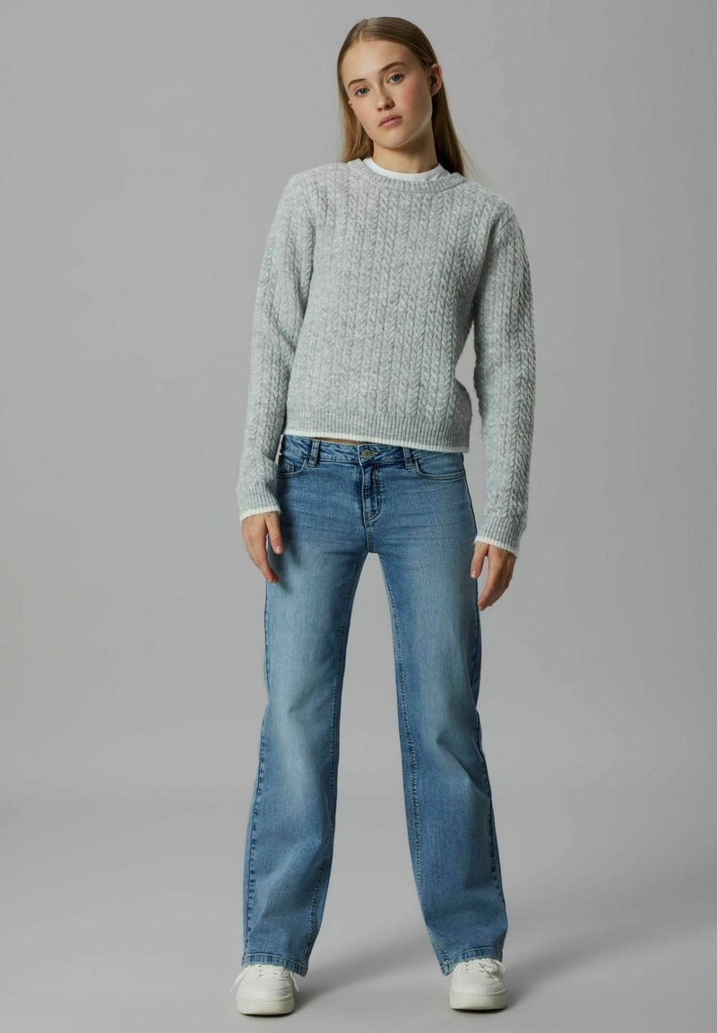 Pelēks adīts džemperis ar ribotiem apkakles malām, kombinēts ar gaiši ziliem, brīva piegriezuma džinsiem. Modelis stāv pret neitrālu fonu.