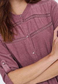 Femme portant une chemise mauve boutonnée avec des insertions en dentelle et des manches retroussées, les bras croisés.