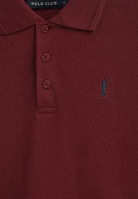 Bordeauxfärgad polo med en strukturerad tyg, som har en treknapplock och en liten blå broderad logotyp av en golfspelare på bröstet.