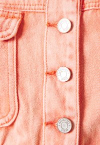 Närbild av persikofärgad denim med tre silverknappar framtill, prydd med kontrasterande orange sömnadsdetaljer.