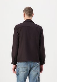 Chemise marron foncé avec col, manches longues et poignets boutonnés, associée à un pantalon cargo en denim bleu clair avec de multiples poches.