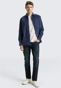 Jonge man in een blauwe overhemd met knopen over een wit t-shirt, donkere jeans en witte sneakers, staande met één hand in zijn zak.