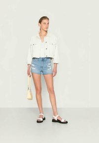 Jaqueta de ganga branca cropped, t-shirt branca, calções de ganga azuis desgastados e sandálias preto e branco. Segurando uma pequena bolsa amarela.