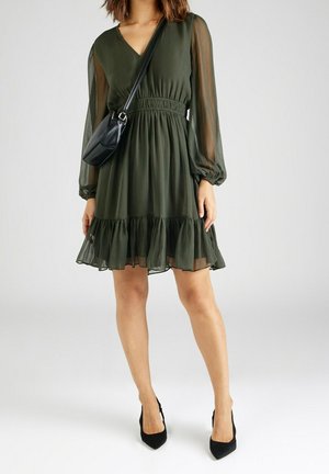 Femme portant une robe longue manches longues vert foncé avec des manches transparentes, un ourlet à volants, un sac bandoulière noir et des talons hauts noirs, debout sur un fond blanc.