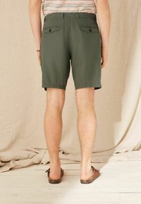 Pantaloni corti verdi realizzati in tessuto leggero, con due tasche posteriori con chiusure a bottone. Indossati con scarpe slip-on marroni.
