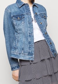 Veste en denim bleu clair avec un col à revers, des poches avant et des fermetures à boutons ; présente une finition douce et texturée.