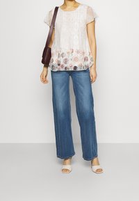 Blusa leggera e trasparente bianca con motivi floreali sull'orlo, maniche corte e bottoni; abbinata a jeans a gamba larga blu e sandali chiari.