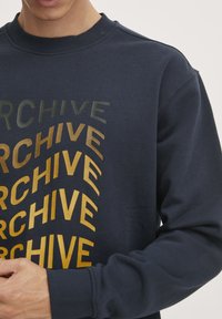 Sudadera azul marino con cuello de canalé, que presenta el texto en negrita y curvado "ARCHIVE" en dorado y gris oscuro en la parte frontal. Textura suave.