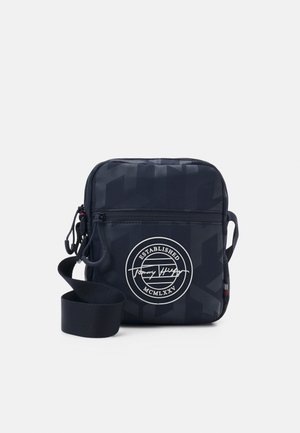 Kompakt marineblå crossbody-taske med justerbar rem, frontlynlåslomme og hvidt rundt logo med teksten "Established MCMLXXV."