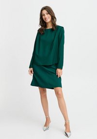Robe deux pièces verte comprenant un haut à manches longues avec un décolleté bateau et une jupe ajustée au genou. Texture lisse et design minimaliste.