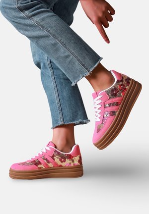 Personne portant des baskets plateforme roses à motif floral avec des lacets blancs et un jean bleu raccourci effiloché, un pied levé.