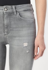 Jean en denim gris à taille haute, avec des accents déchirés et une texture lisse. Comprend une poche avant et un détail de bouton en métal.