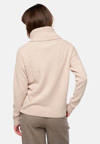 Beige Strickpullover mit hohem, geripptem Kragen und überschnittenen Schultern. Getragen mit strukturierten braunen Hosen. Stoff ähnlich wie Wolle, weiche Textur.