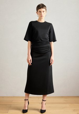 Mustat mekko, jossa on lyhyet, leveät hihat ja ryhdikäs vyötärödetalji. Tasainen kangas, nilkkapituus ja suora siluetti. Yksinkertainen leikkaus. Mustat korkokengät.