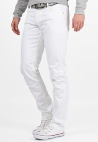 Jean en denim blanc avec des coutures jaunes, coupe slim, et un design de poche avant. Portés avec des baskets blanches avec des rayures colorées.