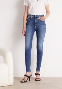 Hnědé džíny s vysokým pasem, slim fit, vyrobené z denimu. Mezi jejich vlastnosti patří vybledlý předek, pět kapes a knoflíkový zip. Nosené s černými podpatky.