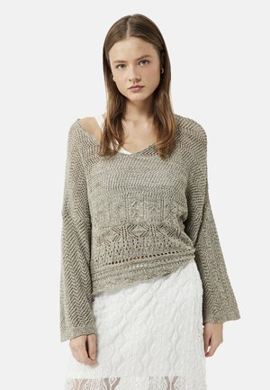 Jonge vrouw met lang bruin haar, die een losgebreide beige trui draagt over een witte top en een witte kanten rok, kijkt naar voren.