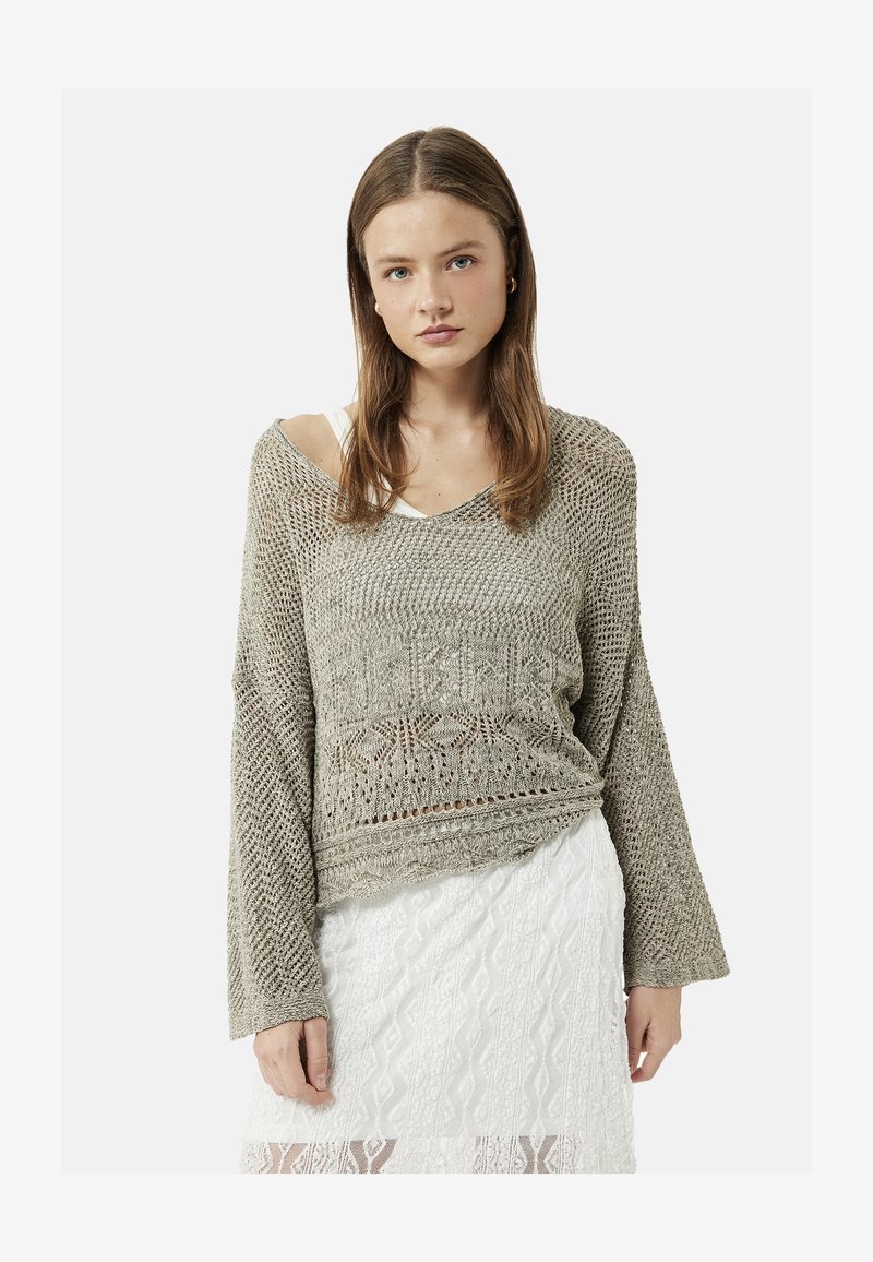 Jeune femme aux longs cheveux bruns portant un pull beige en maille ample par-dessus un haut blanc et une jupe blanche en dentelle, face à l'avant.