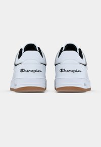 Białe trampki z czarnymi akcentami, wykonane z gładkiej skóry, z zaokrąglonym noskiem, wyściełanym kołnierzem i gumową podeszwą w kolorze gumy. Logo "Champion" po bokach.