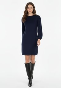Abito blu navy con maniche lunghe, colletto rotondo e tre bottoni decorativi dorati sulla spalla; tessuto morbido con una texture liscia.