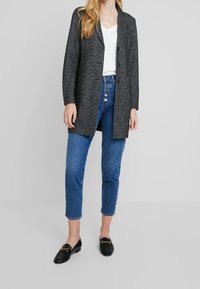 Cappotto in misto lana grigio con colletto a rever, chiusura con un bottone singolo e tasche frontali, abbinato a jeans di denim blu a vita alta e mocassini neri.