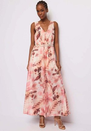 Donna che indossa un abito lungo rosa con fiori, senza maniche e scollo a V, con una sottile cintura e sandali con tacco nude, in piedi davanti a uno sfondo neutro.