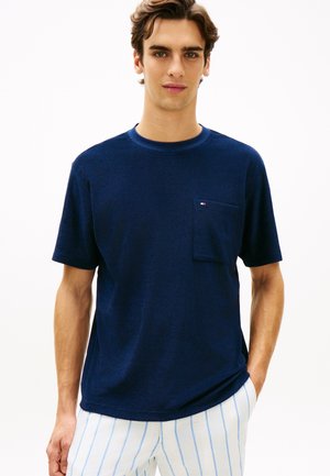 Jonge man die een marineblauw shirt met korte mouwen en borstzak draagt, en witte broek met lichtblauwe verticale strepen.