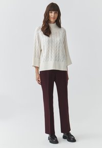 Maglione in lana a maglia cable color crema con colletto alto, abbinato a pantaloni dritti bordeaux e mocassini scuri. Sfondo semplice.