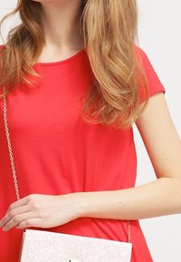 Rotes Kurzarmoberteil aus weichem Stoff, getragen mit einer hellrosa glitzernden Clutch. Das Oberteil hat einen einfachen runden Ausschnitt und eine lockere Passform.