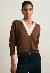 Maglione a maglia marrone con colletto bianco, caratterizzato da un design a V. Abbinato a jeans in denim scuro, con cuciture e tasche visibili.