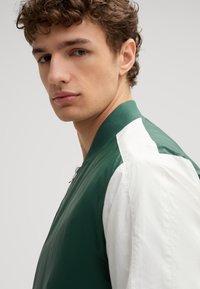 Giacca bomber verde e bianca con chiusura a zip, colletto a costine e design a pannelli a contrasto. Tessuto liscio con materiale traspirante.