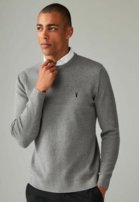 Pull en maille côtelée gris avec un col rond, des manches longues, des poignets ajustés et un logo subtil sur la poitrine. Superposé à un col de chemise blanc.